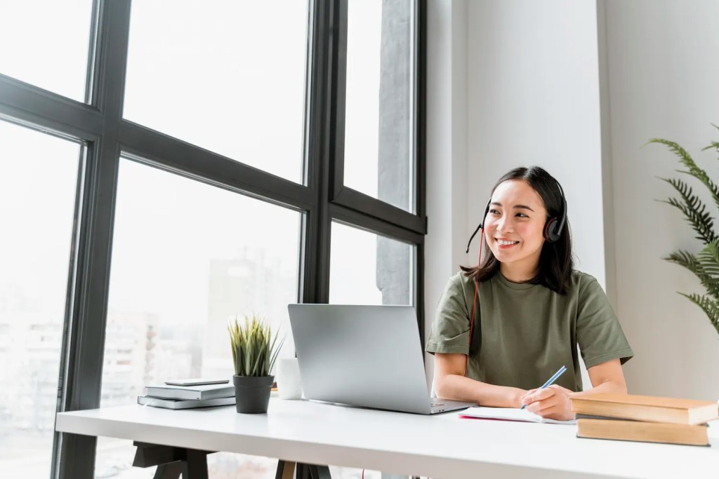 A smiling healthcare support professional from the Philippines wearing a headset and looking out the window while taking notes at her desk, with a laptop and books in front of her. - Platonics LLC