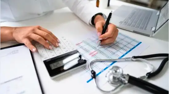 Medical Billing Outsourcing in the Philippines A medical professional's hands use a calculator and pen on a document with a stethoscope and laptop nearby, representing medical billing outsourcing in the Philippines.
